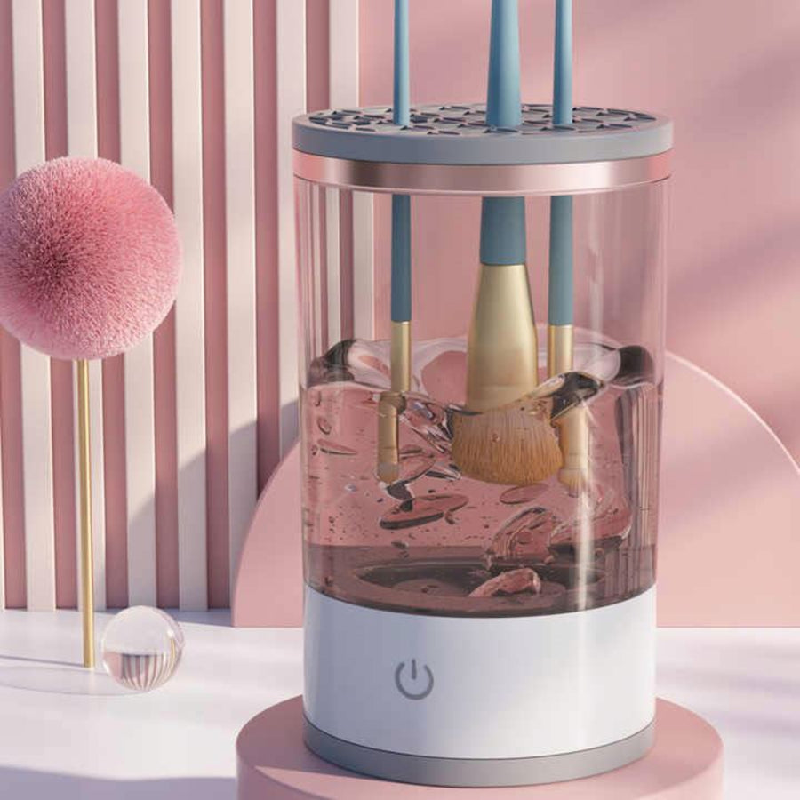 A pink and white electric makeup brush cleaner with a clear cylindrical chamber, shown with several makeup brushes inside. The device sits on a pink circular base against a pink striped background, with a decorative pink pom-pom prop on the left.